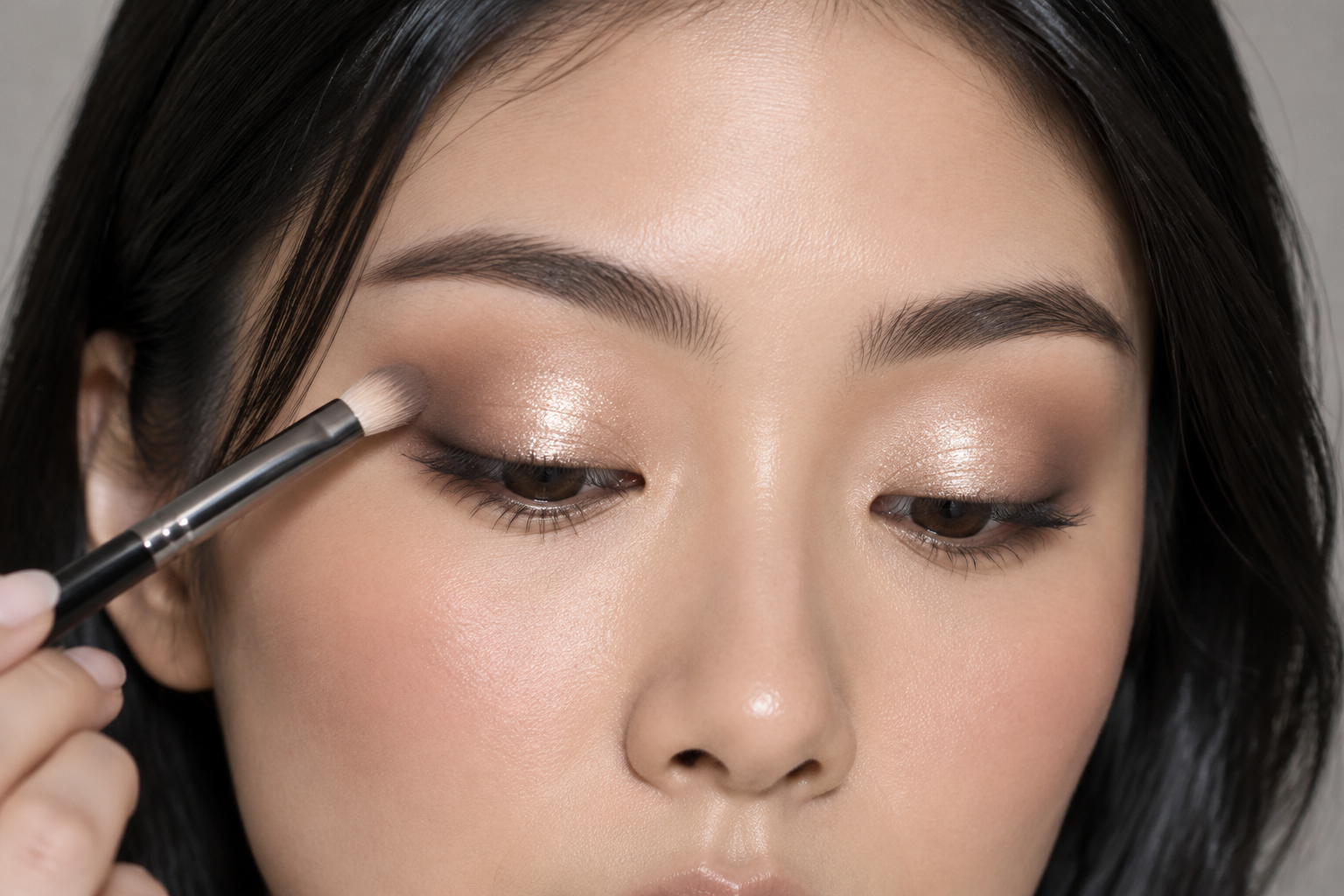 An East Asian woman with lowered eyes holds a small blending brush against the outer edge of champagne shimmer on her eyelid, beside soft matte crease shading.