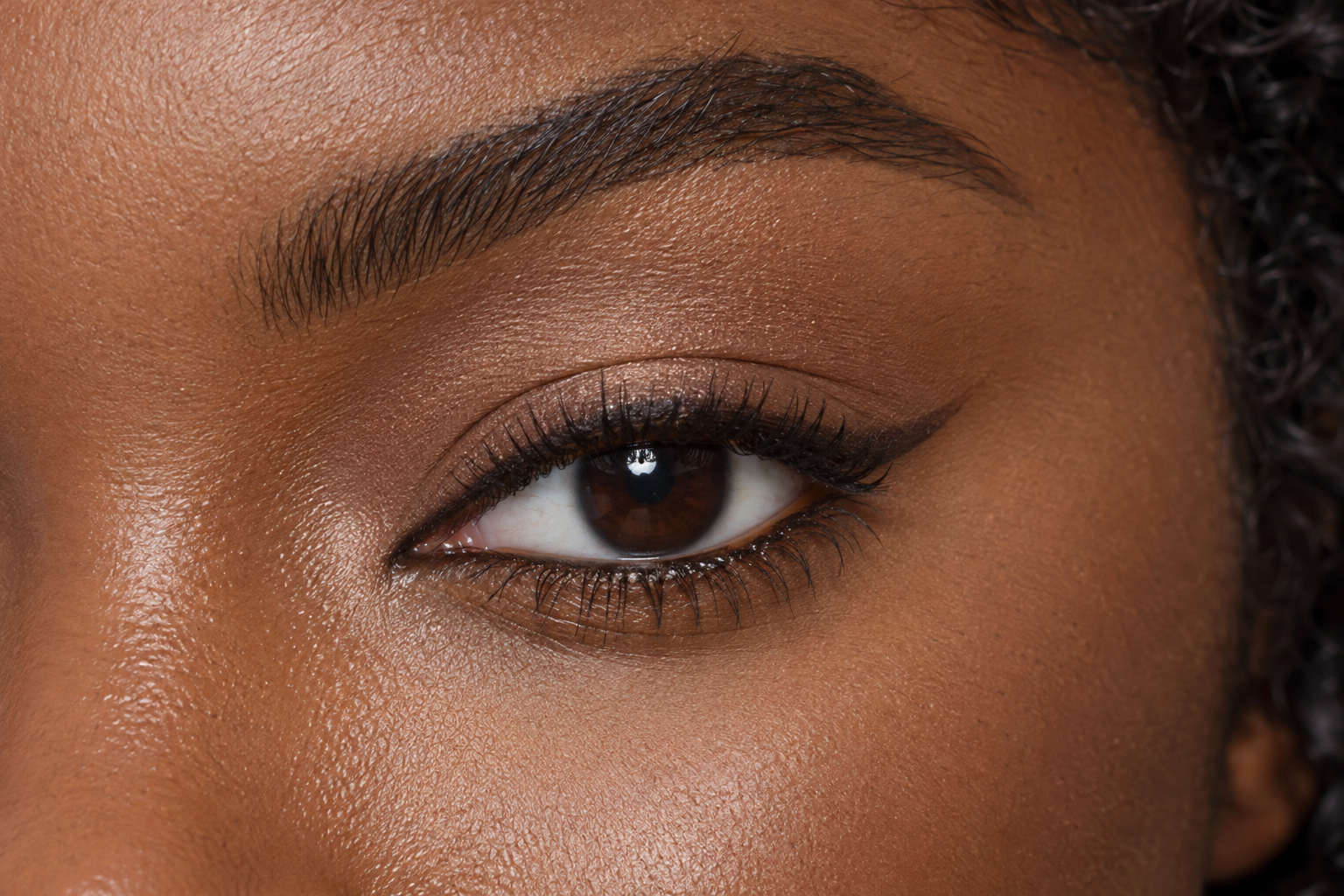 Close view of a hooded eye with narrow espresso liner along the upper lashes and a short outer flick.