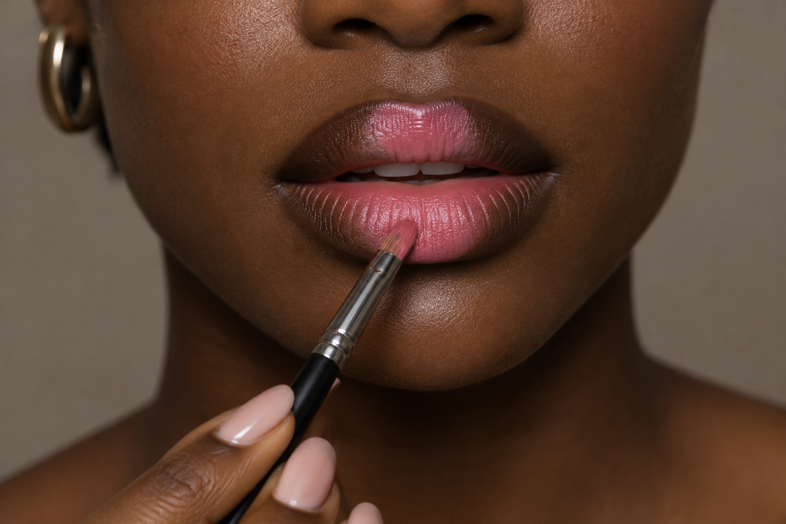 A woman with deep dark skin applies rose-pink lipstick to the center of her lower lip with a small brush, with espresso-brown color visible around both lips.