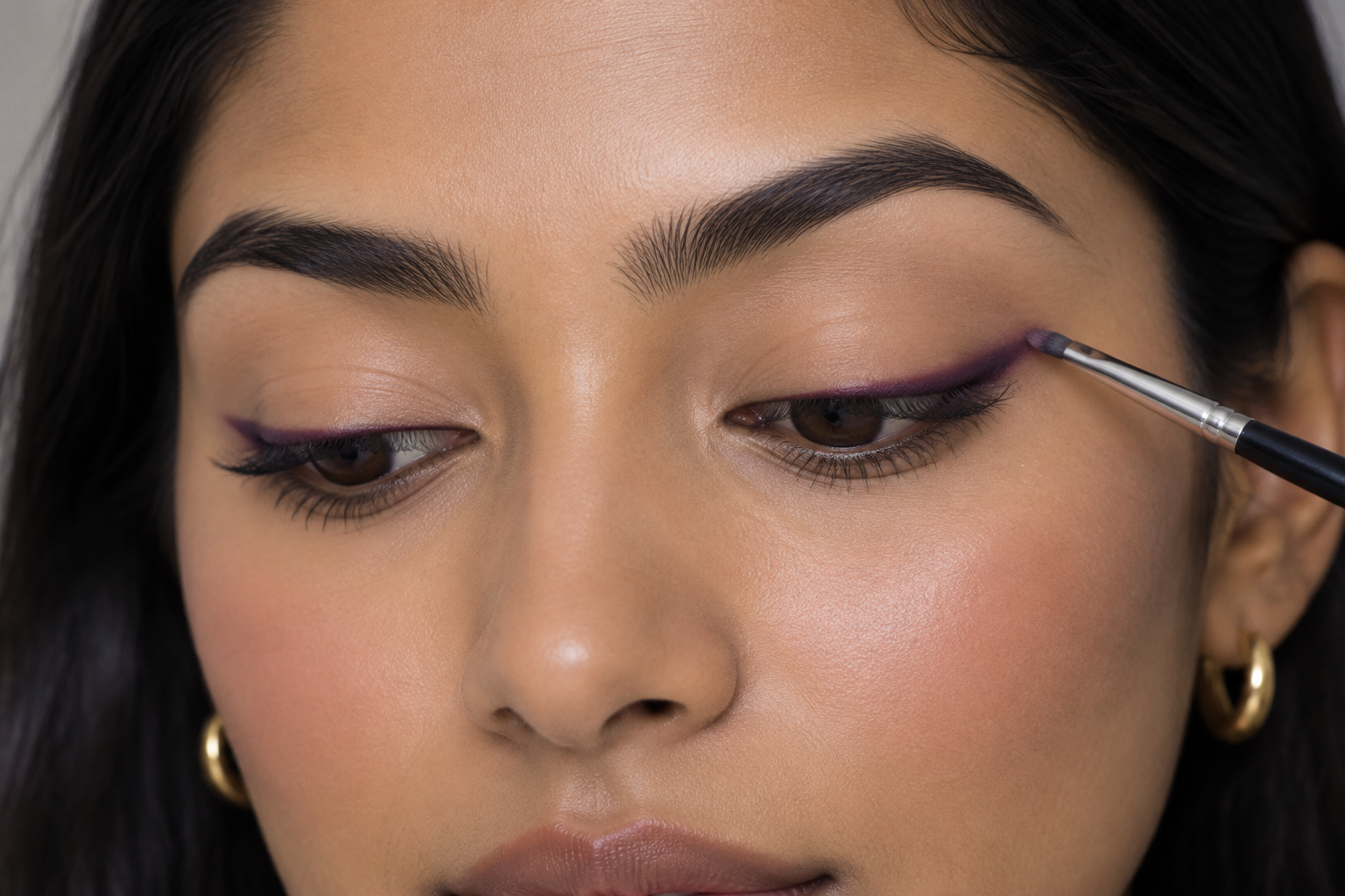South Asian woman with lowered eyes holding a small brush at the upper edge of a narrow plum line along her upper lashes.