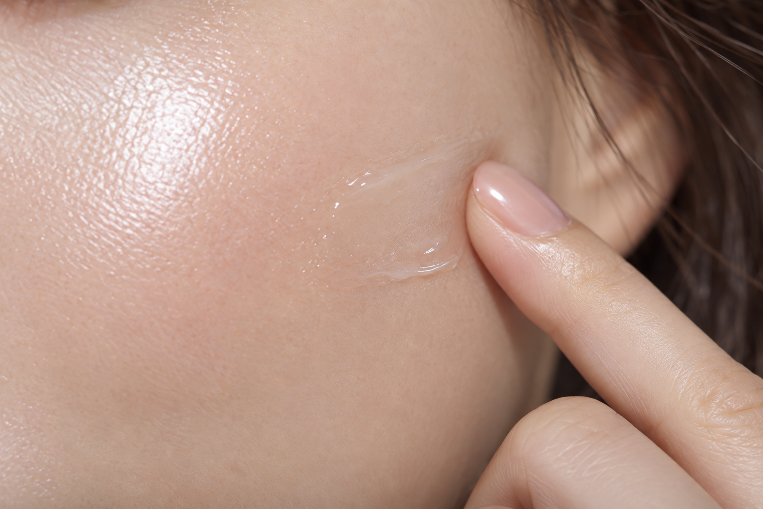 A fingertip lightly touching a cheek, with a small translucent skincare streak at the contact point.